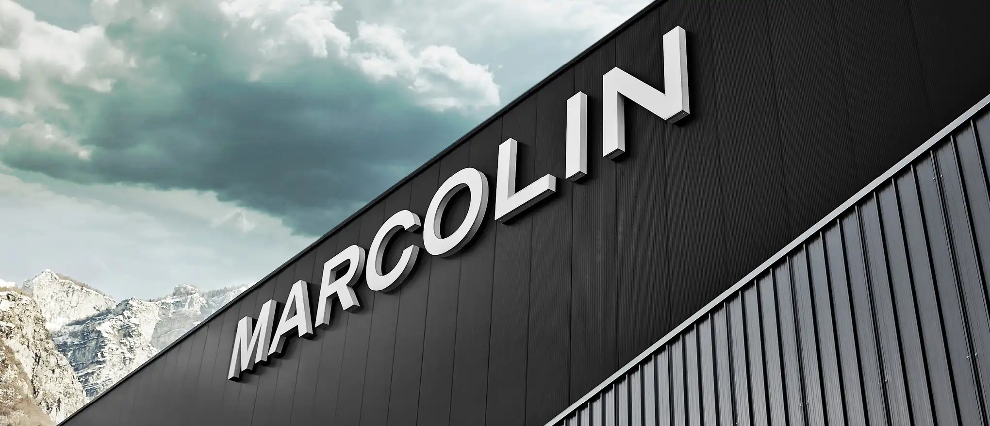 Modern building with 'MARCOLIN' logo, set against mountains and a cloudy sky.